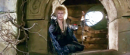David Bowie dressed in a black cape and sparkling fantasy costume with a blonde spiky haircut, sitting in a castle window spinning crystal balls before releasing them onto the wind.