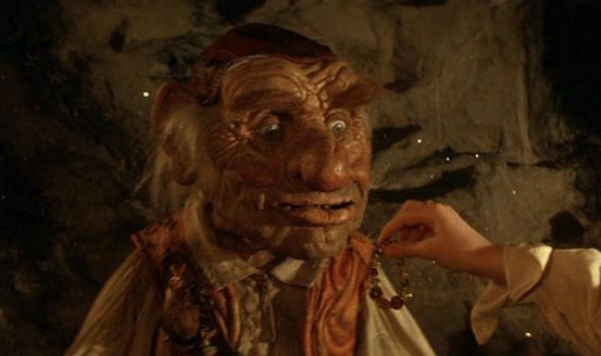A wrinkly-faced dwarf man with a red leather skull-cap, orange waistcoast and white smock shirt. He looks amazed as a girl hands him a plastic bracelet.