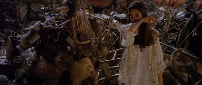 A teenage girl in a cream shirt and waistcoat stands in a junkyard. She is talking to a wrinkled old woman laden with junk.
