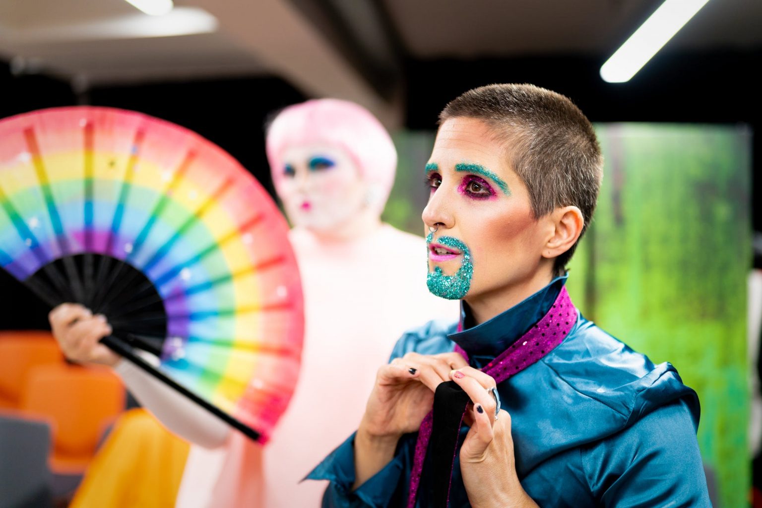 A drag king with a neat blue beard and a blue jacket, adjusts their purple tie.